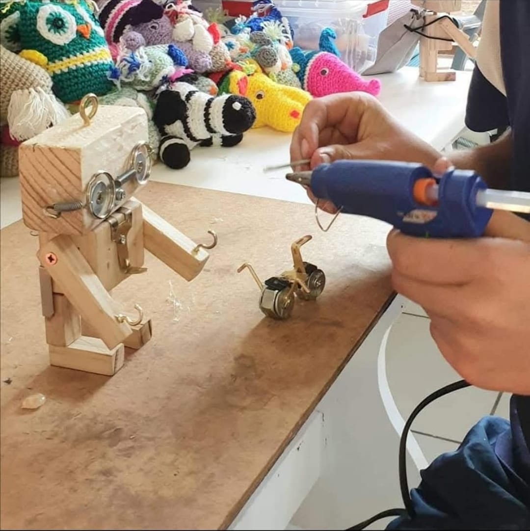 Hands using a glue gun beside a wooden robot and a tiny metal bicycle on a work board