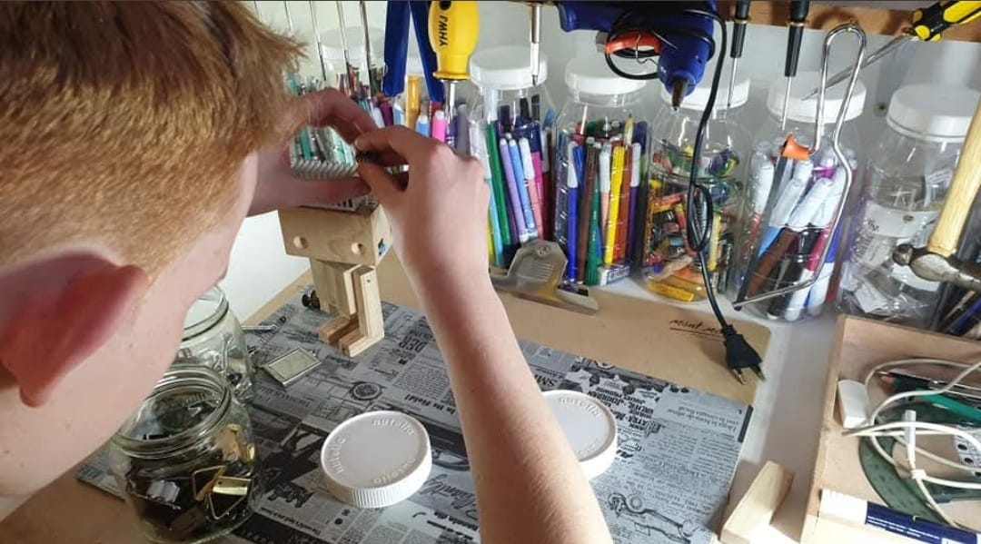 A young maker assembling a small wooden figure at a bench under a rack of tools and pens