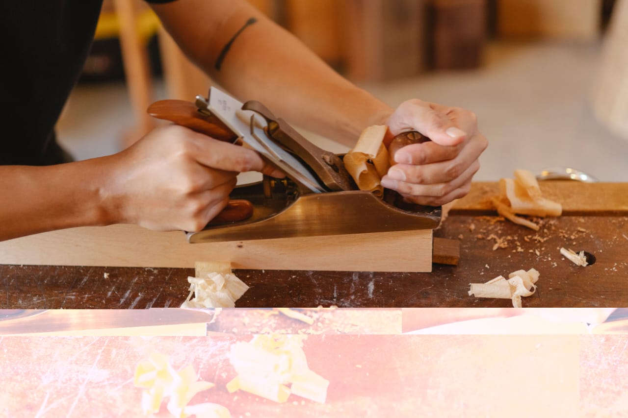 Hands guiding a hand plane along a piece of wood, with curls of shavings on the bench