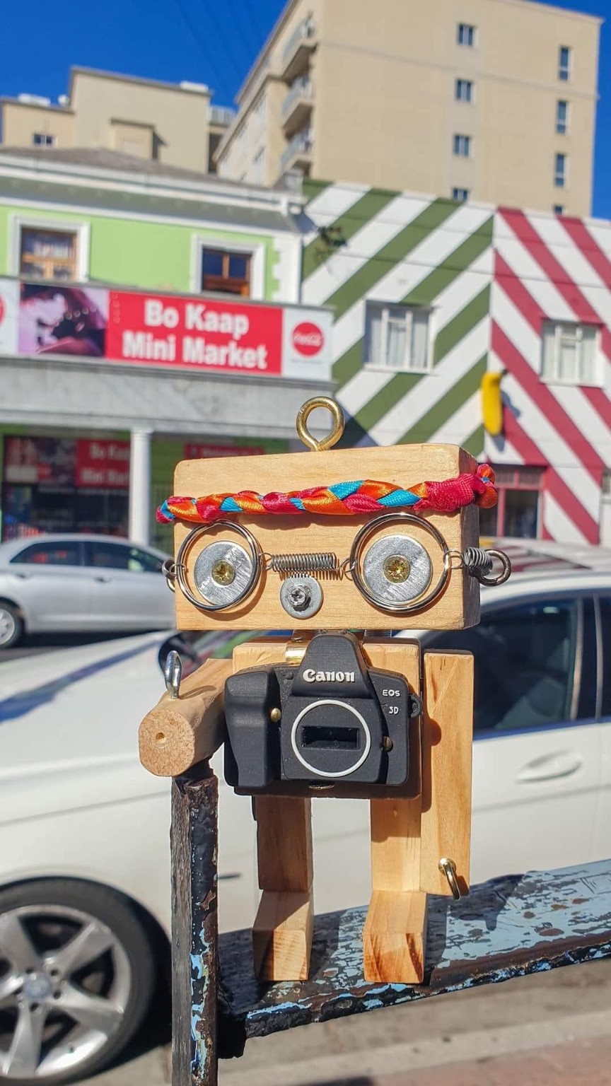 A wooden robot with a braided headband and camera, photographed outdoors in front of a Bo-Kaap street scene