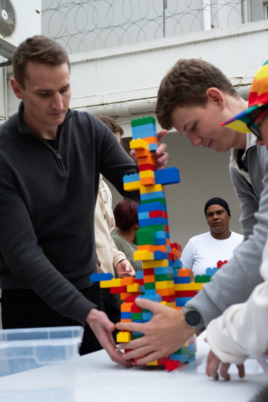 Colleagues steadying a tall brick tower together as they build it on a table outdoors.