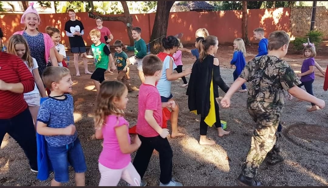 A large group of children in capes playing together outdoors under trees