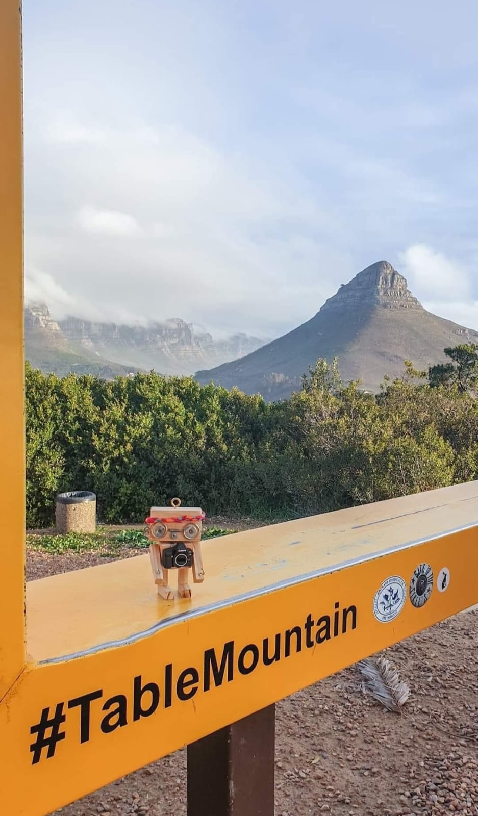 A small wooden camera robot standing on a yellow Table Mountain sign with Lion's Head behind