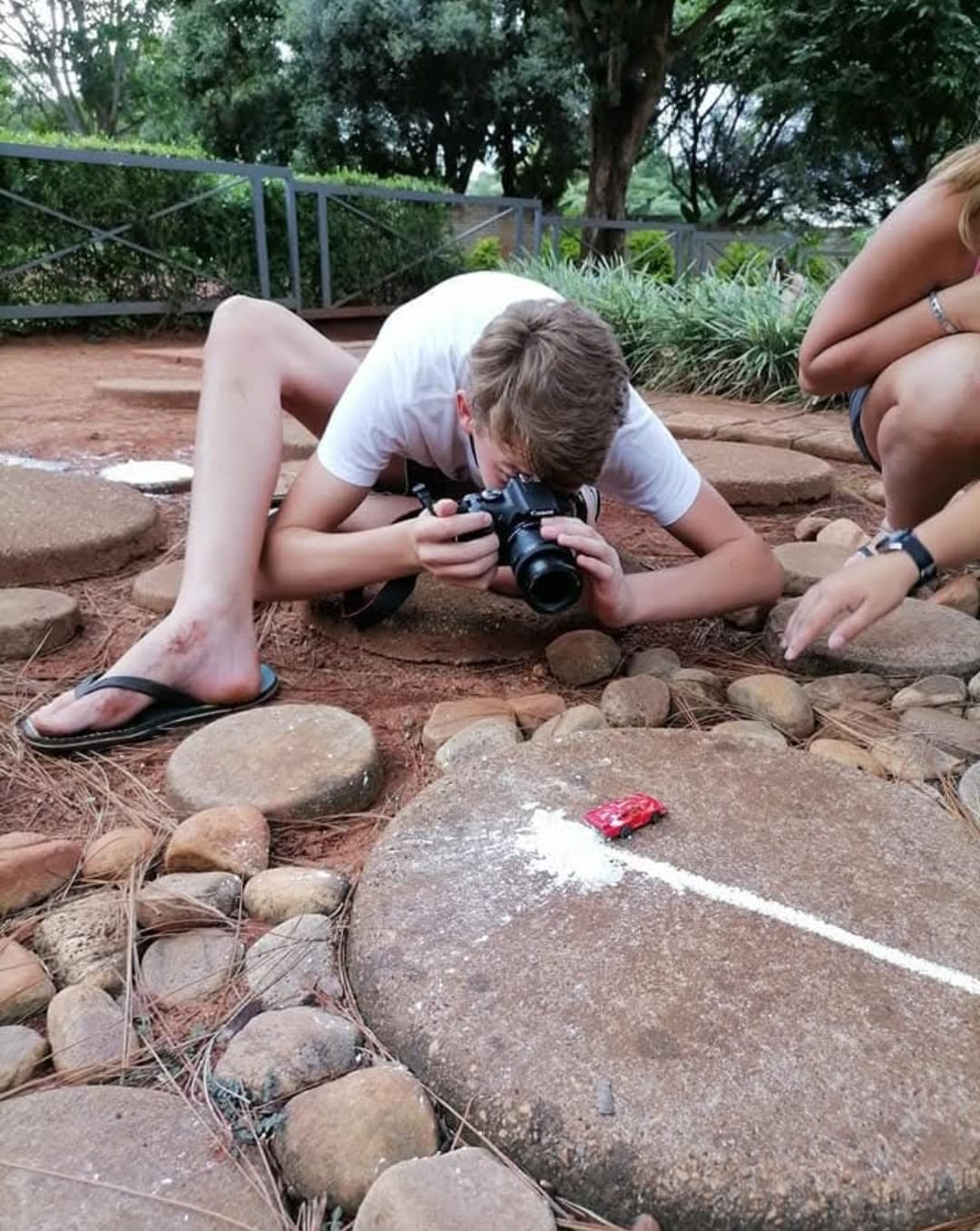 A boy lying on the ground photographing a small red toy car on a stone with a chalk track