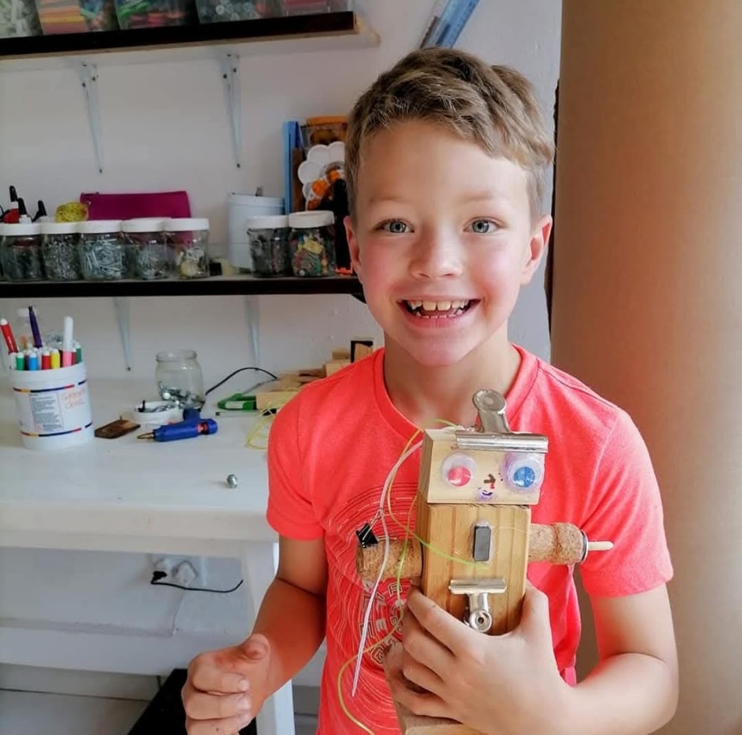 A grinning boy holding up the wooden robot he built, workshop shelves of jars behind him