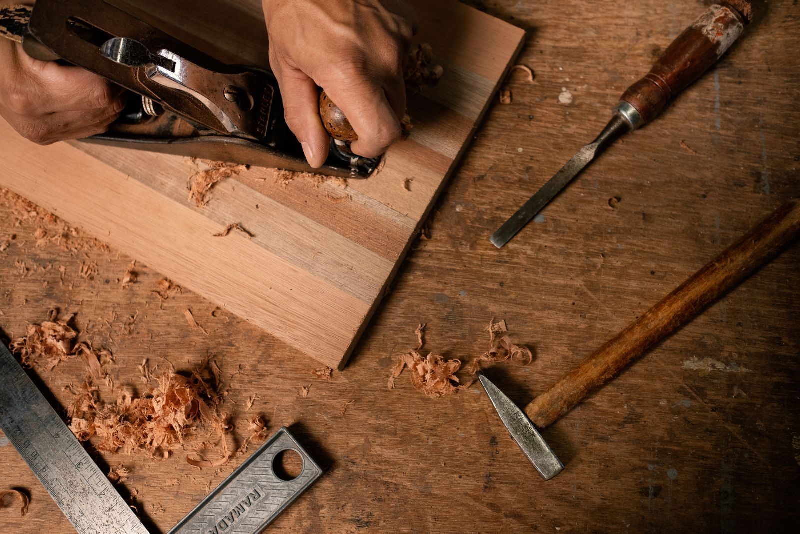 Hands running a hand plane along a plank, with chisels and shavings on the bench