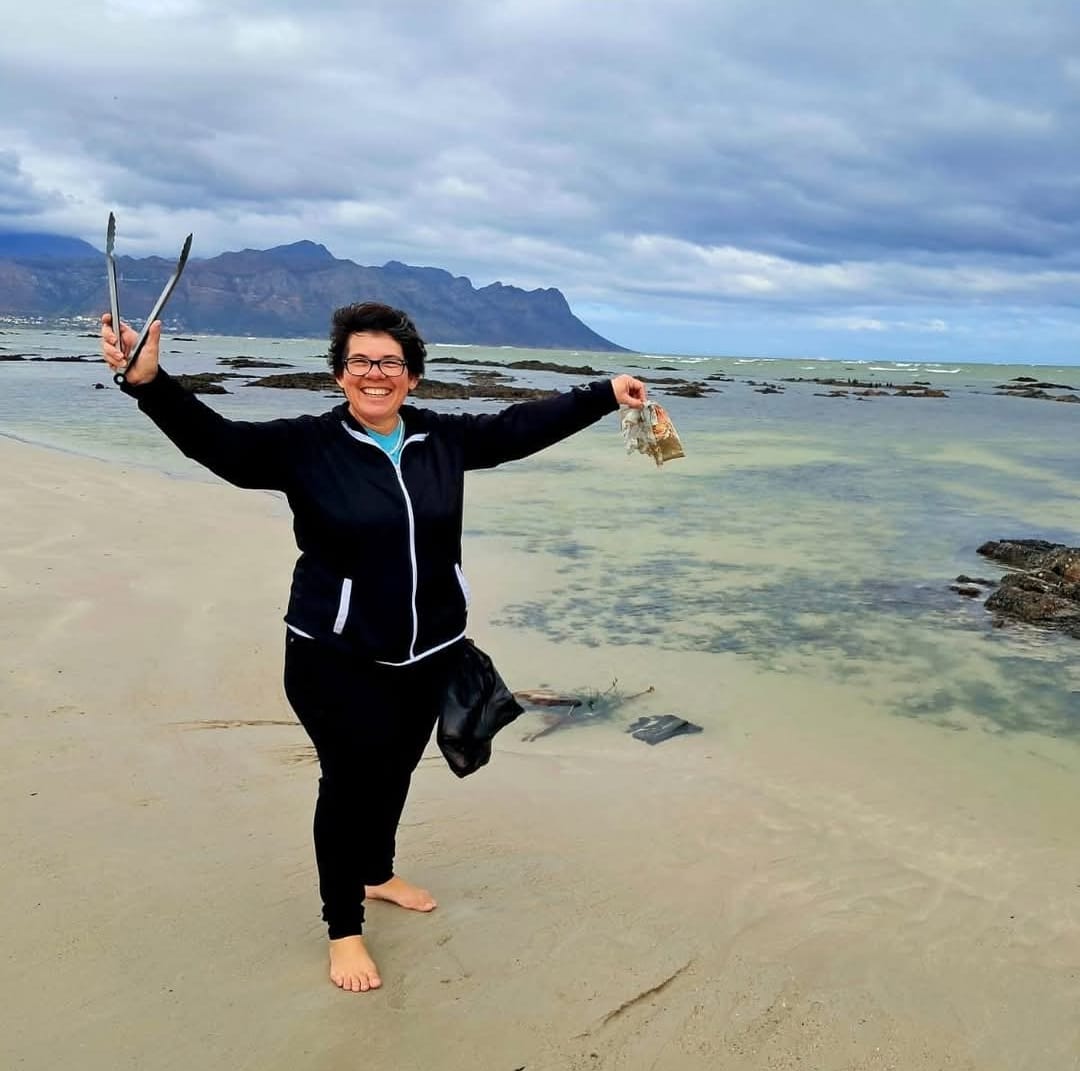 Sonia standing on a beach with Table Mountain in the background, holding tongs in one hand and a collection bag in the other